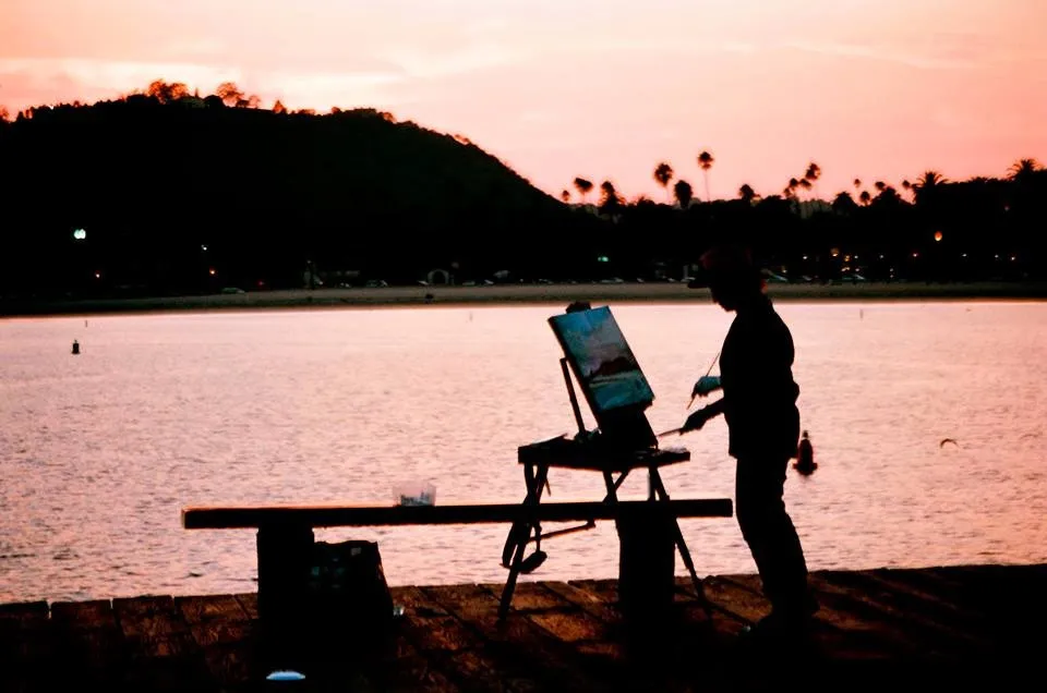 Michele Zuzalek painting outdoors at an easel at sunset, seen in silhouette by the water