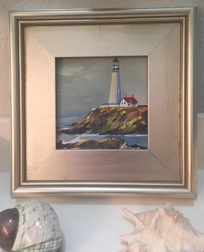 A coastal lighthouse painting in a wide silver frame, shown with seashells